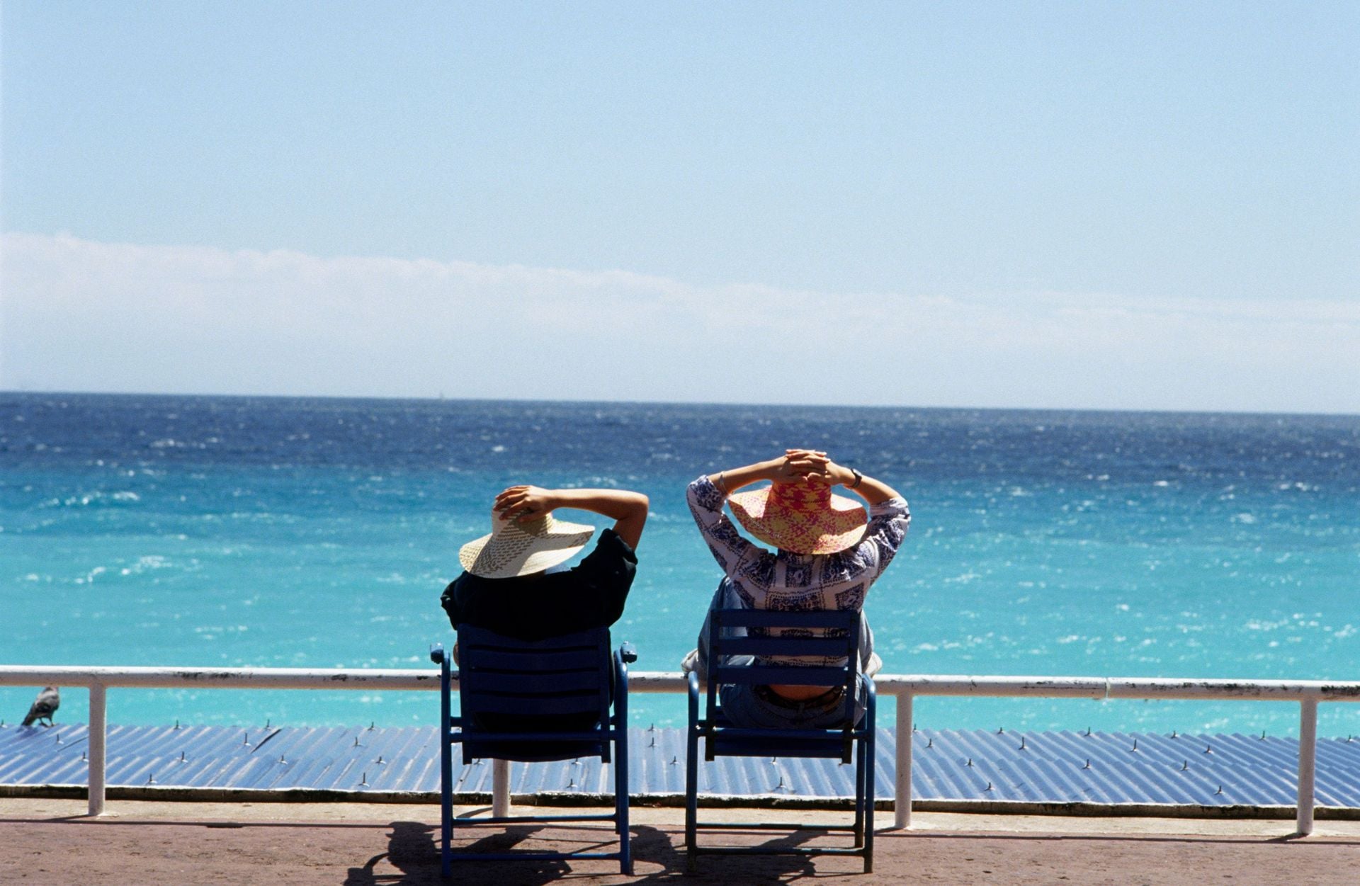 France, Nice, Promenade des Anglais, two women in straw hats sitting on deck chairs facing the sea, rear view.