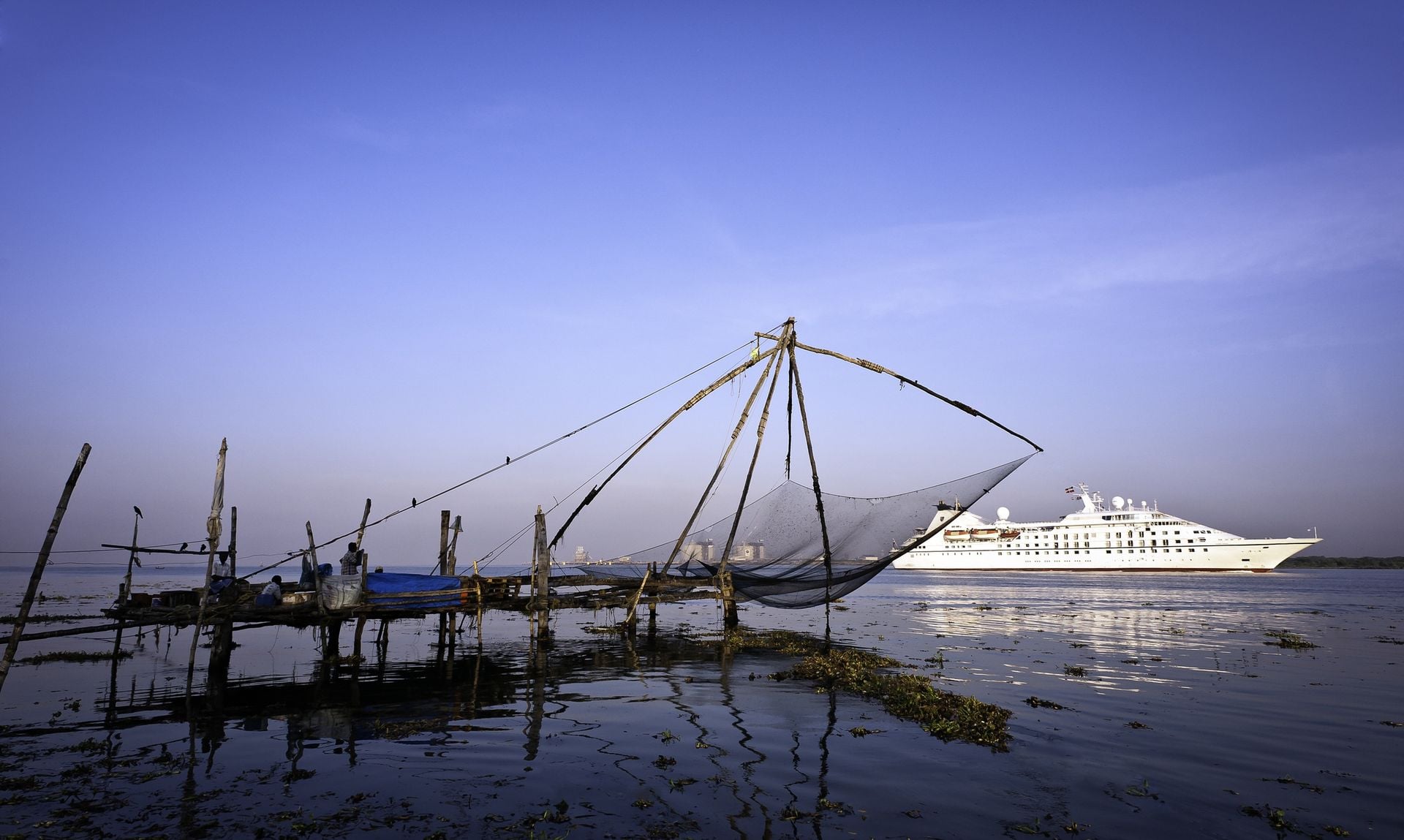 Fort Kochi, Kerala, India. A passenger ship enters Fort Kochi harbour as a Chinese Fishing Net rests in the calm waters of the Arabian Sea at sunset in Kerala, south India.