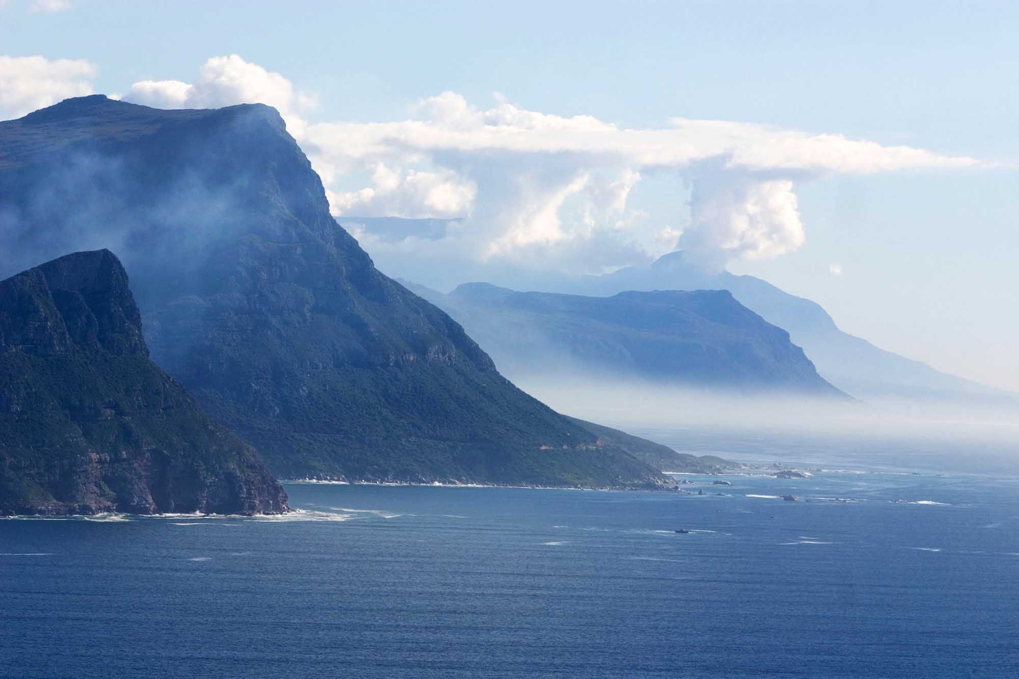 Fog hanging just above the deep blue sea entering the land at Cape of Good Hope, South Africa, with column-like clouds hanging over the mountains