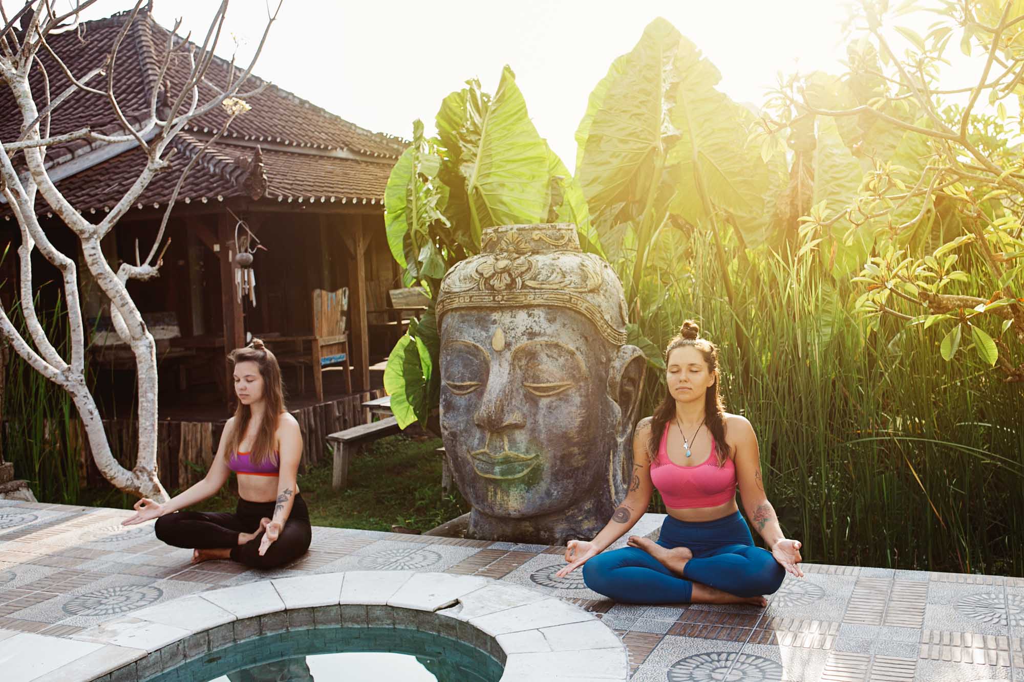 Young women practicing yoga during luxury yoga retreat in Asia, Bali, meditation, relaxation, getting fit, enlightening, green grass jungle background, buddha statue